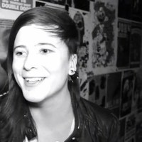 A black and white photo of Stef at an art show; it is a candid photo of them smiling during an interview. They are a white person with freckles and long straight hair that is cropped short on one side.